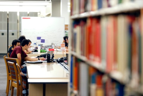 Students sit at tables surround by bookshelves in the library with books and laptops
