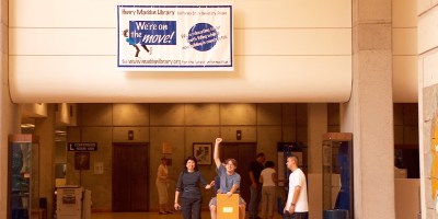 Library workers take a book cart for a ride while moving books to the south wing during construction. Sign above them reads, "We're on the move!"