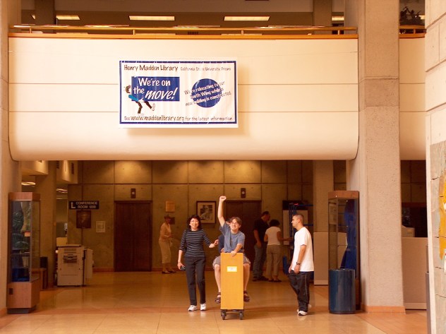 Library workers take a book cart for a ride while moving books to the south wing during construction. Sign above them reads, "We're on the move!"
