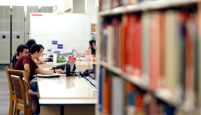 students study with computers at library tables with blurred book shelves in the foreground