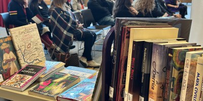 A collection of books sits in the foreground and a group of people listen in the background.